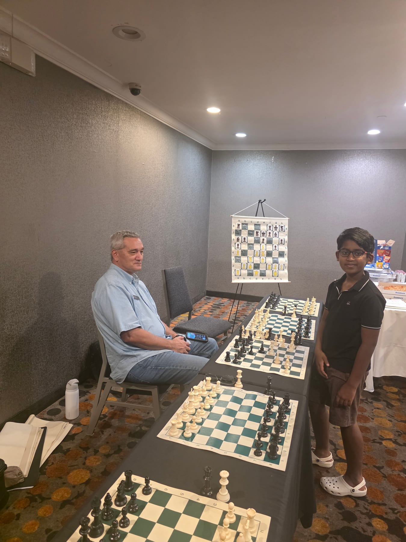 An adult and a young player beside a row of chessboards.